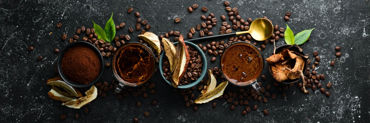 Small bowls of ground coffee, cacao and dried mushrooms arranged in a row on dark slate with scattered coffee beans.
